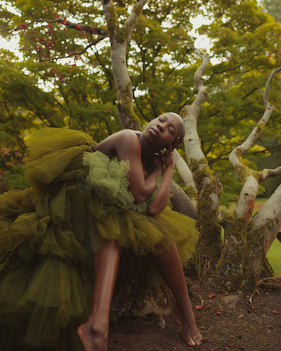 Dancer in a treed area in a yellow-green tutu with bared arms, shoulders and legs below the knee
